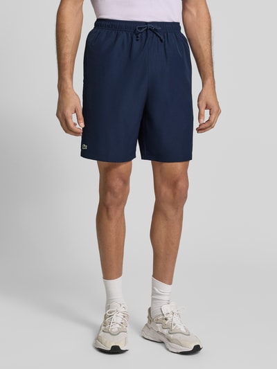 Männermodel trägt marineblaue Relaxed Fit Shorts mit Logo-Patch von Lacoste Sport, glatte Struktur, Ganzkörperansicht von vorne.