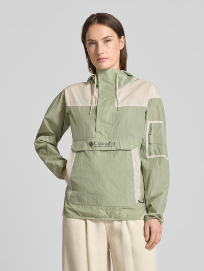 Frauenmodel trägt Regular Fit Windbreaker mit Logo-Print Modell 'Challenger' von Columbia in Hellgrün und Beige, strukturierte Oberfläche, Känguru-Tasche, Kapuze mit Tunnelzug, elastische Abschlüsse, ärmellange 57 cm, Vorderansicht, Nahaufnahme Oberkörper.