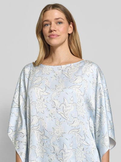 Modelka nosi Loose Fit Poncho-Bluse mit Allover-Muster od s.Oliver BLACK LABEL, efekt: wzór na całej powierzchni, krój loose fit, dekolt w łódkę, rękawy nietoperzowe, wykończenie materiału: tkanina, materiał: czysta wiskoza, jasnoniebieskie i kremowe kolory, lekka tekstura, zbliżenie na górną część ciała.