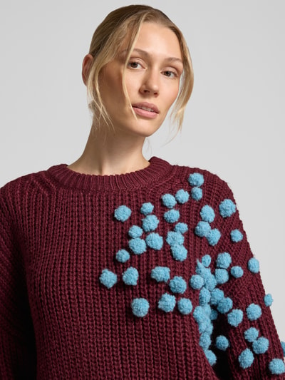 Frauenmodel trägt Relaxed Fit Pullover mit Woll-Anteil Modell 'KOSTAR' von Essentiel Antwerp in Dunkelrot mit blauen Pompons, Nahaufnahme Oberkörper. Damen Pullover von Essentiel Antwerp.