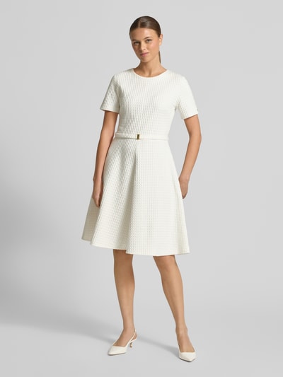 Kobiece modelka nosi Knielangens Kleid aus Baumwoll-Mix mit Strukturmuster od Marc Cain, mieszanka bawełny, linia A, krótkie rękawy, okrągły dekolt, detal z logo, odpinany pasek z materiału, długość do kolan, sukienka taliowana, kolor jasny, fakturowana powierzchnia, widok całej sylwetki z przodu.