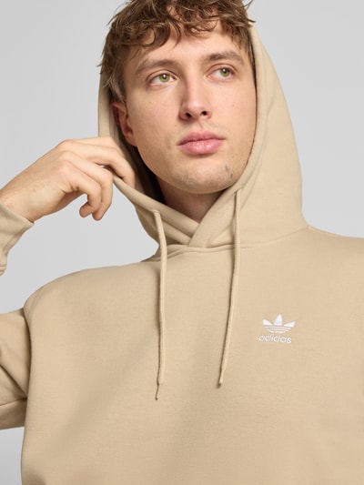Męski model nosi Hoodie mit Känguru-Tasche od adidas Originals, mieszanka bawełny, krój regular fit, wyhaftowane logo, beżowy, zbliżenie na górę.