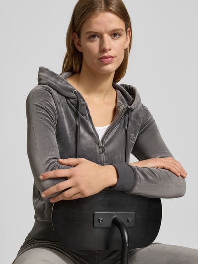 Frauenmodel trägt Sweatjacke mit Label-Stitching Modell 'ROBERTSON' von Juicy Couture, grauer Material-Mix mit Stretch-Anteil, Regular Fit, Kapuze mit Tunnelzug, Reißverschluss und Känguru-Tasche, Nahaufnahme Oberkörper.