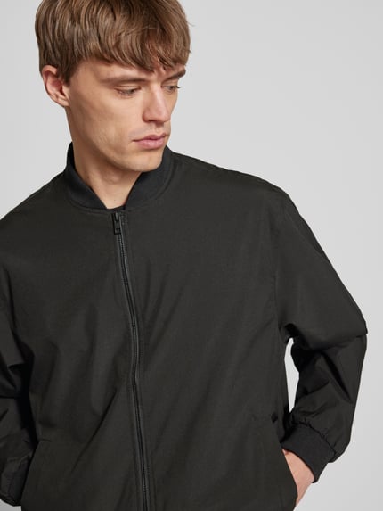 Męski model nosi Bomberjacke mit seitlichen Eingrifftaschen Modell 'CHARGE' von Jack & Jones w czerni, krój straight fit, stójka, zamek błyskawiczny, boczne kieszenie, prążkowane wykończenia, długość tyłu w rozmiarze M: 66 cm, szerokość ramion w rozmiarze M: 54 cm, szerokość klatki piersiowej w rozmiarze M: 65 cm, długość rękawa w rozmiarze M: 64 cm, gładka tekstura, widok z przodu, zbliżenie na górną część ciała.