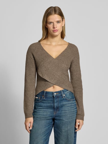 Frauenmodel trägt Pullover mit Wickeloptik von PIECES in Braun mit Rippstrickstruktur zu blauen Jeans, Ganzkörperansicht von vorne.