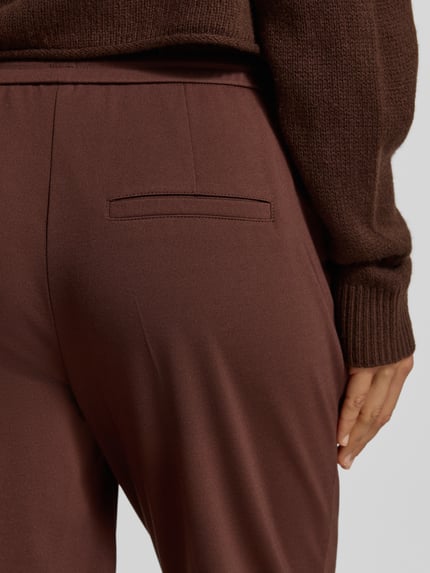 Frauenmodel trägt Straight Fit Hose aus Viskose-Mix mit Bundfalten von Luisa Cerano in Braun, Detailaufnahme Nahaufnahme Hüfte. Elastischer Bund sichtbar, glatte Textur, Bügelfalten, verkürzte Passform, eleganter Look.