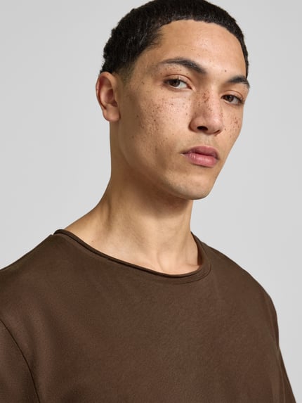 Męski model nosi T-Shirt mit Rollsaum od Filippa K z bawełny, prosty krój, okrągły dekolt, zrolowany brzeg, długość tyłu 70 cm, zbliżenie na górną część ciała.