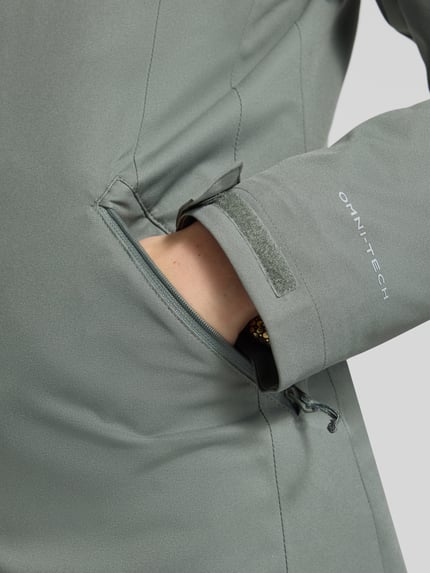 Frauenmodel trägt Regular Fit Jacke mit Kapuze Modell 'Pulaski' von Columbia in Grau, strukturierte Oberfläche, Detailaufnahme, taillierte Passform, Eingrifftasche sichtbar, KI-modifiziertes Bild.