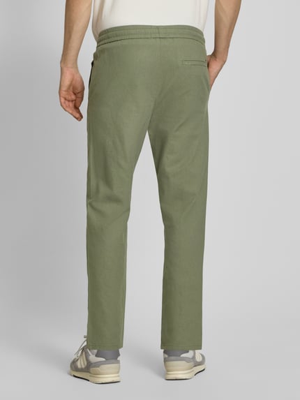 Męski model nosi Tapered Leinenhose aus Baumwoll-Leinen-Mix Modell 'LINUS' od Only & Sons, kolor zielony melanż, krój tapered fit, elastyczny pas, kieszenie boczne i tylne, materiał bawełniano-lniany, widok całej sylwetki z tyłu.