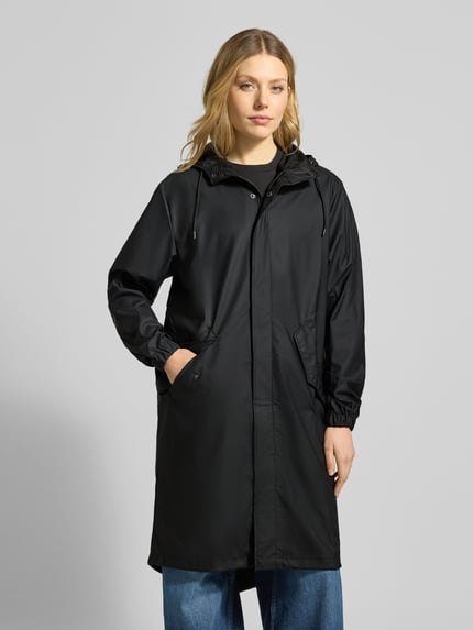 Frauenmodel trägt schwarzen Regular Fit Parker mit Kapuze Modell 'Fishtail' von Rains mit Pattentaschen und elastischen Ärmelabschlüssen, Ganzkörperansicht von vorne. Rückenlänge 104 cm, Schulterbreite 45 cm, Brustweite 55 cm, Ärmellänge 65 cm, Glanztextur.
