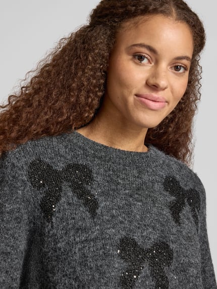 Frauenmodel trägt Pullover mit Glitzer-Schleifenprint von ONLY, melierte Strickoptik, Motiv-Print, Effektgarn, Nahaufnahme Oberkörper.
