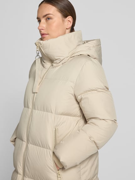 Frauenmodel trägt Regular Fit Daunenmantel mit abnehmbaren Rucksackriemen von Marc O'Polo in Beige, große Steppnähte, glatte Textur, Nahaufnahme Oberkörper. Damen Daunenmantel von Marc O'Polo.