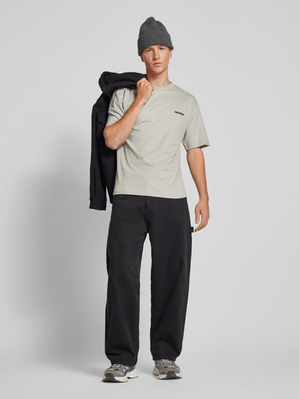 Männermodel trägt Relaxed Fit T-Shirt aus reiner Baumwolle von REVIEW in Beige mit Logo-Stickerei, schwarze Hose, Ganzkörperansicht von vorne.