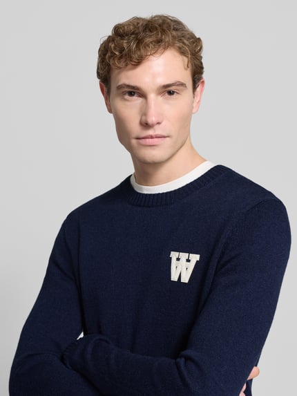 Männermodel trägt Strickpullover aus Lammwolle Modell 'Tay' von Wood Wood in Dunkelblau mit Label-Stitching, Rundhalsausschnitt, Regular-Fit, Nahaufnahme Oberkörper. Rückenlänge bei Größe M 72 cm, Ärmellänge 68 cm.