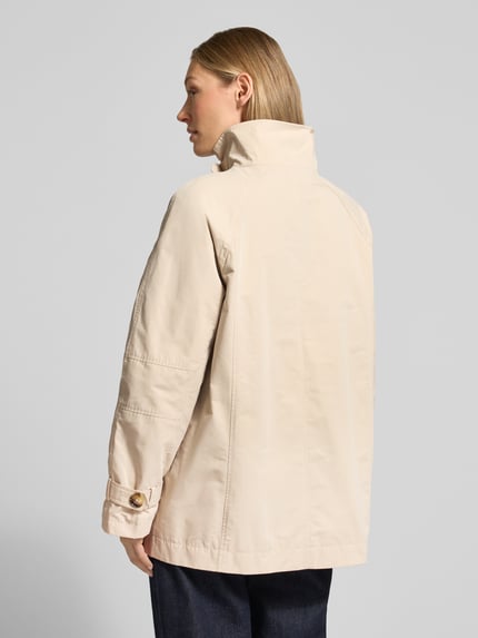 Frauenmodel trägt Trenchcoat mit Stehkragen und Ärmelriegeln von Fuchs Schmitt, beigefarben, strukturierte Baumwolle, Ganzkörperansicht Rückansicht.