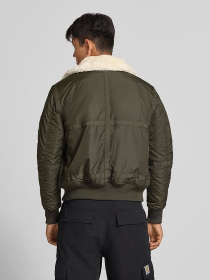 Männermodel trägt Bomberjacke mit Label-Patch und Kentkragen von Alpha Industries in Olivgrün, Teddyfellkragen, Rückansicht, Ganzkörperansicht Rückansicht. Regular Fit, Reißverschluss, gerippte Abschlüsse, Reißverschlusstasche am Ärmel, Eingrifftaschen mit Druckknopfverschluss, Rückenlänge 65 cm, Schulterbreite 45 cm, Brustweite 56 cm, Ärmellänge 65 cm.