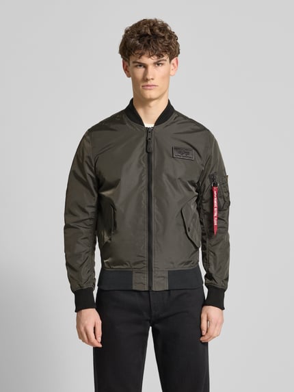 Męski model nosi Regular Fit Bomberjacke mit Logo-Patch od Alpha Industries w kolorze oliwkowym, stójka, łatka z logo, kieszenie, zamek, kieszeń na rękawie, długość tyłu w rozmiarze M: 65 cm, szerokość ramion w rozmiarze M: 45 cm, szerokość klatki piersiowej w rozmiarze M: 54 cm, długość rękawa w rozmiarze M: 69 cm, widok całej sylwetki z przodu.