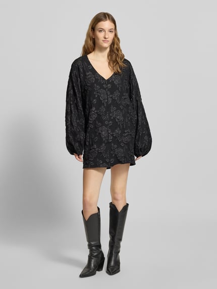 Vrouwenmodel draagt Minikleid mit V-Ausschnitt van Gina Tricot, los vallend, V-hals, sierbies, elastische mouwuiteinden, zwart met bloemenpatroon, volledige lichaamsweergave van voren