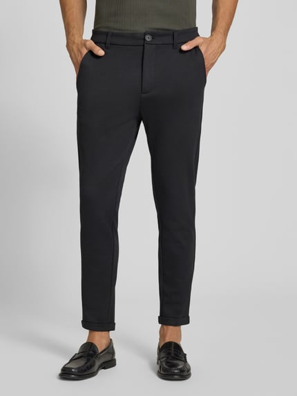 Męski model nosi Slim Fit Hose mit Viskose-Anteil in verkürzter Passform von Lindbergh, czarny, mieszanka wiskozy, wpuszczane i tylne kieszenie, zapięcie na guzik i zamek błyskawiczny, szerokość w rozmiarze M: 42 cm, długość wewnętrzna nogawki: 72 cm, krój slim fit, skrócony krój, materiał o gładkiej fakturze, spodnie materiałowe, Ganzkörperansicht z przodu.