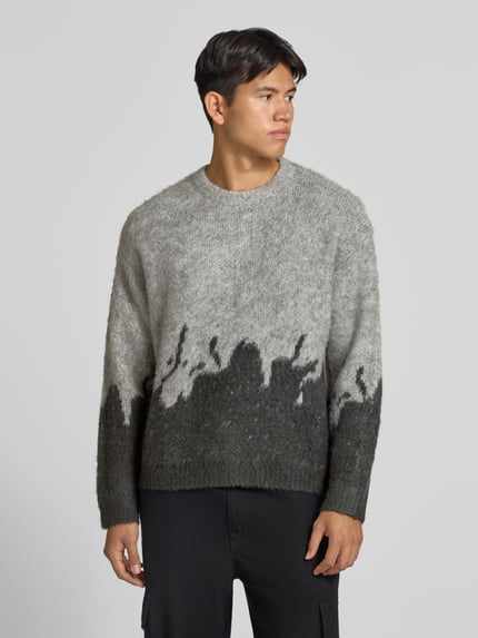 Männermodel trägt Strickpullover mit gerippten Abschlüssen von ANOTHER COTTON LAB in Grau, lockerer Schnitt, melierte Textur, Nahaufnahme Oberkörper. Elastischer Material-Mix mit Lanawoll-Anteil, Rundhalsausschnitt, überschnittene Ärmel, Rückenlänge 62 cm, Ärmellänge 67 cm.