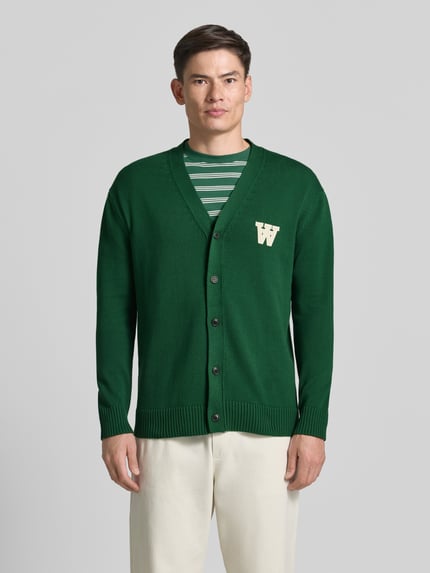 Männermodel trägt grüne Strickjacke mit V-Ausschnitt Modell 'Tay' von Wood Wood aus Reine Baumwolle, Gerader-Schnitt, Ganzkörperansicht von vorne.