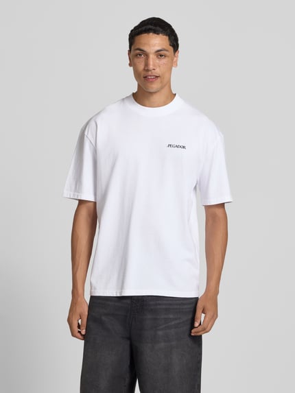 Männermodel trägt wit oversized T-shirt van Pegador, puur katoen, model 'Miles' met labelprint, ronde hals, heren, ruglengte maat M 70 cm, Ganzkörperansicht van voren.