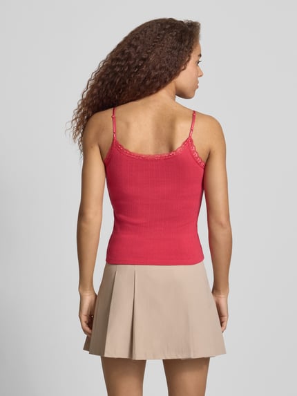 Kobiecy model nosi Slim Fit Top mit Spitzenbesatz od REVIEW, czysta bawełna, krój slim fit, ramiączka spaghetti, ozdobne kokardki, długość tyłu w rozmiarze S: 38 cm, jasnoczerwony, beżowa plisowana spódnica, pełny widok sylwetki z tyłu.