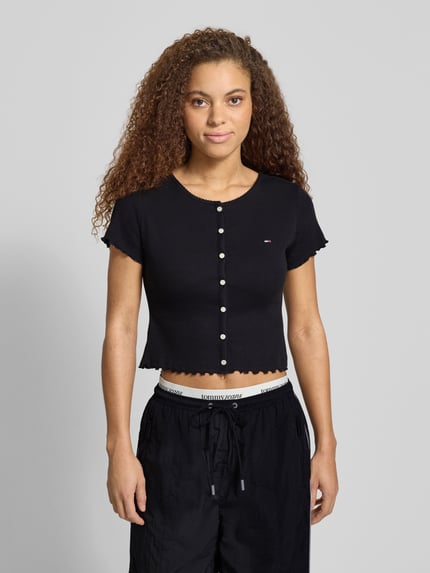 Vrouwenmodel draagt Slim Fit T-Shirt aus Baumwoll-Mix van Tommy Jeans in uni zwart met logo-stitching, frill afwerking, ribstructuur, korte mouw, U-boothals, Ganzkörperaanzicht van voren.