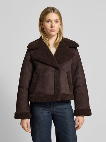 Kobieta modelka nosi Regular Fit Kurtkę w imitacji skóry z podszewką z futerka, model 'HARPER' od Vero Moda, efekt: jednolity kolor, krój regular fit, kołnierz z połami, zapięcie na zatrzask, długi rękaw, wpuszczane kieszenie boczne, wykończenie materiału: podszewka z futerka, kolor ciemnobrązowy, widok na cały przód.
