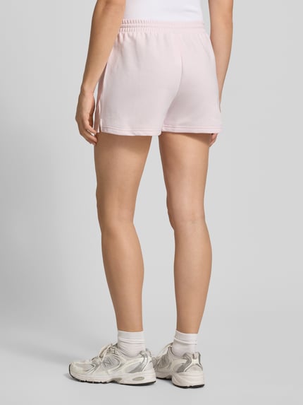 Frauenmodel trägt Straight Fit Shorts mit Tunnelzug Modell 'French' von New Balance aus Baumwoll-Mix in zartem Rosa, Ganzkörperansicht Rückansicht.