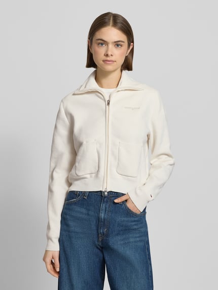 Frauenmodel trägt Slim Fit Strickjacke mit aufgesetzten Taschen von Tommy Jeans in Optik: Unifarben, Strickoptik, Label-Schriftzug, Reißverschluss, Stehkragen, langarm, gerippte Abschlüsse, Materialverarbeitung: Strick, Ganzkörperansicht von vorne.