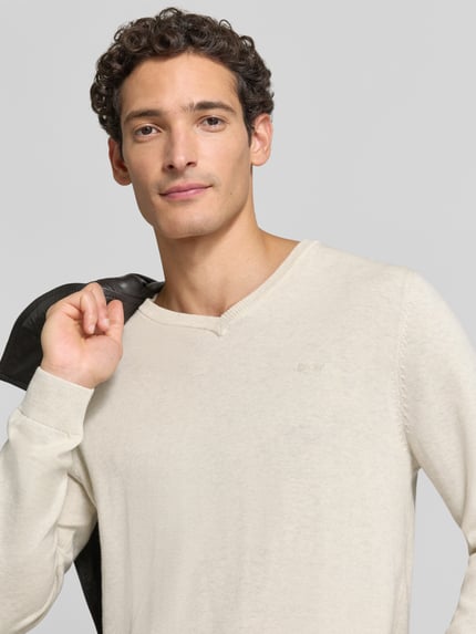 Männermodel trägt Pullover mit Logo-Detail von S.OLIVER CASUAL in Beige, unifarben, V-Ausschnitt, Feinstrick, Nahaufnahme Oberkörper.