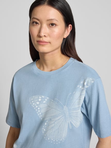 Vrouwenmodel draagt Relaxed Fit T-shirt met motiefprint van Jake*s Casual in lichtblauw, puur katoen, ronde hals, statementprint. Nahaufnahme Oberkörper. Ruglengte bij maat S: 53 cm.