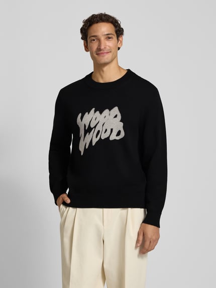 Męski model nosi sweter z dzianiny z bawełny, krój regular fit, nadruk z logo Wood Wood, prążkowane wykończenia, model 'Cloud', długość tyłu 68 cm, długość rękawów 67 cm, widok całej sylwetki z przodu.