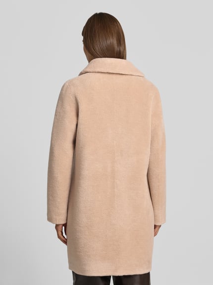 Frauenmodel trägt Wendemantel in Woll-Lammfell-Optik Modell 'VENUS' von OAKWOOD, Beige, lockere Passform, Reverskragen, Eingrifftaschen, Ganzkörperansicht Rückansicht. Rückenlänge 89 cm, Schulterbreite 44 cm, Brustweite 50 cm, Ärmellänge 62 cm.