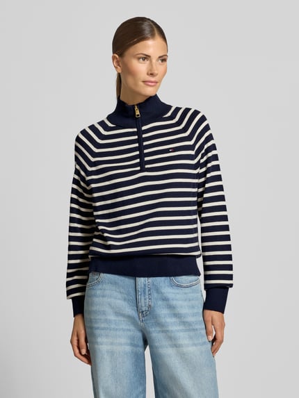 Frauenmodel trägt Pullover mit Troyerkragen von T. HILFIGER SPW., gestreiftes Strickmuster, Reißverschluss, Logo-Detail, Ganzkörperansicht von vorne.