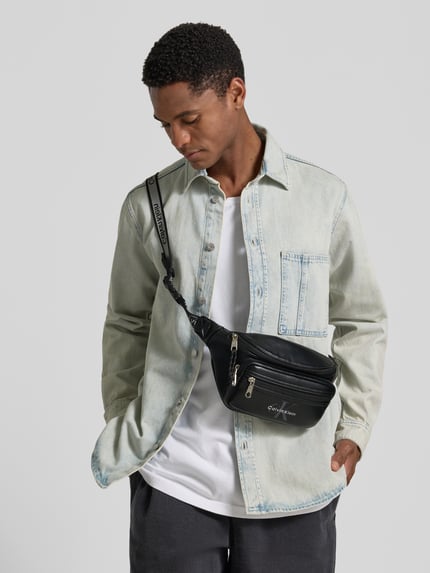 Männermodel trägt schwarze Umhängetasche mit Logo-Prägung Modell 'BOLD LOGO WEBBING WAISTBA' von CK Calvin Klein, Herren, Nahaufnahme Oberkörper.