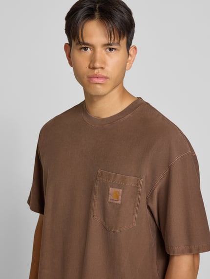 Männermodel trägt Regular Fit T-Shirt aus reiner Baumwolle Modell 'Loose Pocket' von Carhartt Work In Progress in Braun mit Brusttasche, Nahaufnahme Oberkörper.