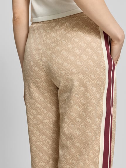 Frauenmodel trägt Wide Fit Sweatpants mit Allover-Logo-Print Modell 'Jacquard' von Guess Activewear, beige mit roten Streifen, Detailaufnahme Rückansicht. Material-Mix mit Baumwoll-Anteil, elastischer Bund, Eingrifftaschen, gemusterte Textur.
