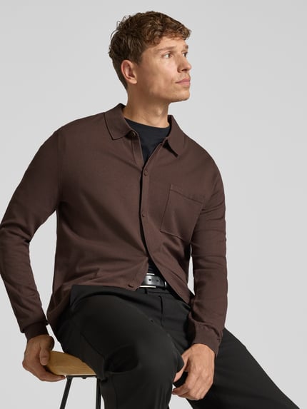 Männermodel trägt Cardigan mit Brusttasche Modell 'San Jordan-CV' von HUGO in Dunkelbraun, Feinstrick, unifarben, Struktur, Longline, Ganzkörperansicht von vorne.