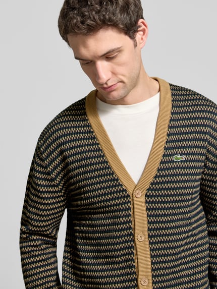 Männermodel trägt Cardigan mit Logo-Applikation von Lacoste, mehrfarbiges geometrisches Allover-Muster in Strickoptik, Nahaufnahme Oberkörper.
