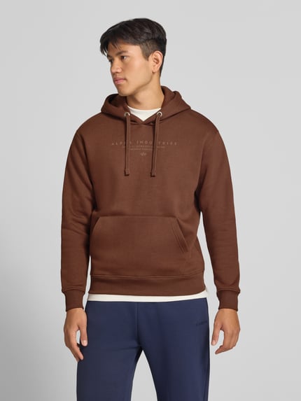Männermodel trägt Relaxed Fit Hoodie aus Baumwoll-Mix mit Label-Detail von Alpha Industries, kieszeń kangurka, tunel, elastyczne wykończenia, Długość tyłu w rozmiarze M: 69 cm, Długość rękawów w rozmiarze M: 62 cm, mieszanka bawełny, brązowy, Ganzkörperansicht von vorne.
