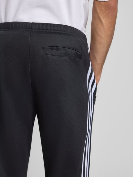 Mannenmodel draagt adidas Originals Trainingshose mit Label-Stitching Modell 'BECKENBAUER', zwart met witte strepen, elastische tailleband, regular fit, katoenmix, achterzak, detailopname heup.