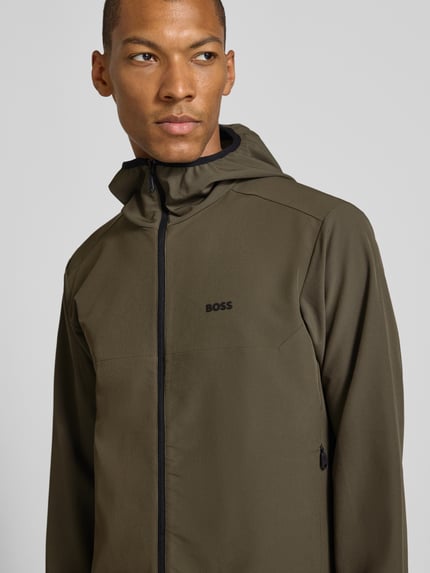 Männermodel trägt Slim Fit Trainingsjacke mit Label-Patch Modell 'CommuterPro' von BOSS für BOSS Green in Oliv mit glatter Struktur, Nahaufnahme Oberkörper.