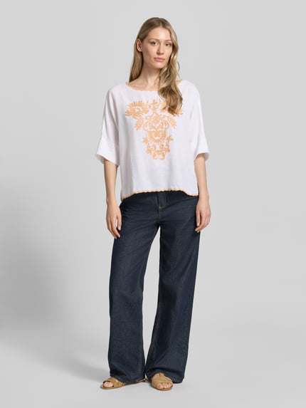Vrouwenmodel draagt Blusenshirt mit Leinen-Anteil und Motiv-Stitching van Christian Berg Woman, wit met oranje motief, viscosemix met linnen, loose fit, boothals met contrasterende rand, ruglengte maat 36: 55 cm, mouwlengte maat 36: 34 cm, ganzlichaamsaanzicht van voren.