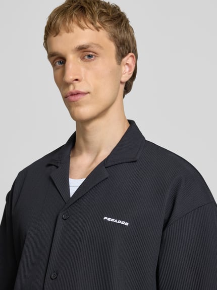 Männermodel trägt Relaxed Fit Freizeithemd mit Reverskragen Modell 'Troy' von Pegador in Schwarz mit Strukturmuster, Logo-Stitching, Knopfleiste, Nahaufnahme Oberkörper.