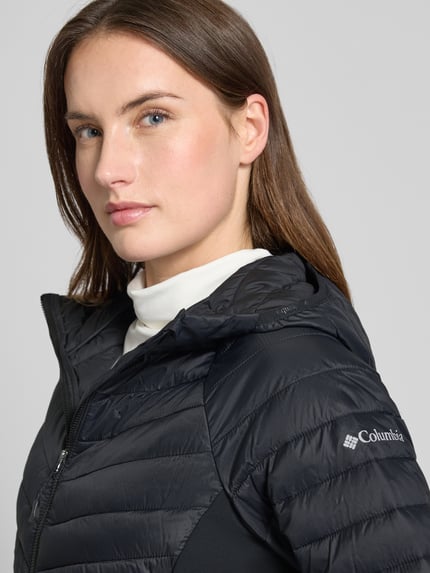 Kobiece modelka nosi Slim Fit Steppjacke mit Reißverschluss Modell 'Omni-Shield' von Columbia, czarny kolor, elastyczny materiał, krój slim fit, kaptur, kieszenie na zamek, elastyczne mankiety, długość tyłu w rozmiarze S: 65 cm, szerokość ramion: 44 cm, szerokość klatki piersiowej: 45 cm, długość rękawa: 60 cm, zbliżenie na górną część sylwetki.
