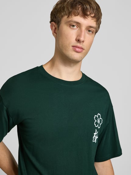 Männermodel trägt Relaxed Fit T-Shirt aus reiner Baumwolle Modell "DYLAN" von JACK & JONES in Dunkelgrün mit Print-Detail, Nahaufnahme Oberkörper.