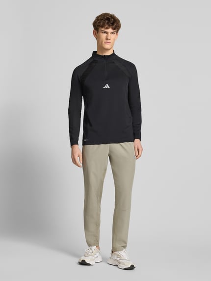 Męski model nosi Sweatshirt mit Stehkragen od adidas Training, czarny, krój slim fit, materiał z rozciągliwością, nadruk logo, długość tyłu 71 cm, długość rękawa 77 cm, widok całej sylwetki z przodu.