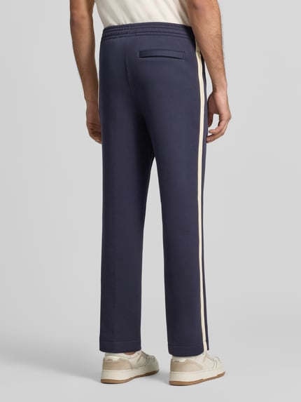 Männermodel trägt Regular Fit Jogpants aus Baumwoll-Mix von Lacoste in Dunkelblau mit cremefarbenem Seitenstreifen, Ganzkörperansicht Rückansicht. Kontext: Herren Weite Hose von Lacoste.